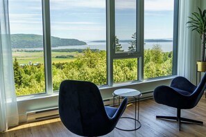 Habitación con amplios ventanales que ofrecen vistas a un lago y follaje. Se observan dos sillas modernas y una pequeña mesa.