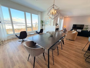 A modern living room with a dining area, large windows, a geometric light fixture, and a flat-screen TV.