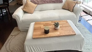 Una sala de estar moderna con sofá, reposapiés y mesa de centro. Hay un ventilador de techo, una ventana con vistas y una puerta que da a otra habitación.