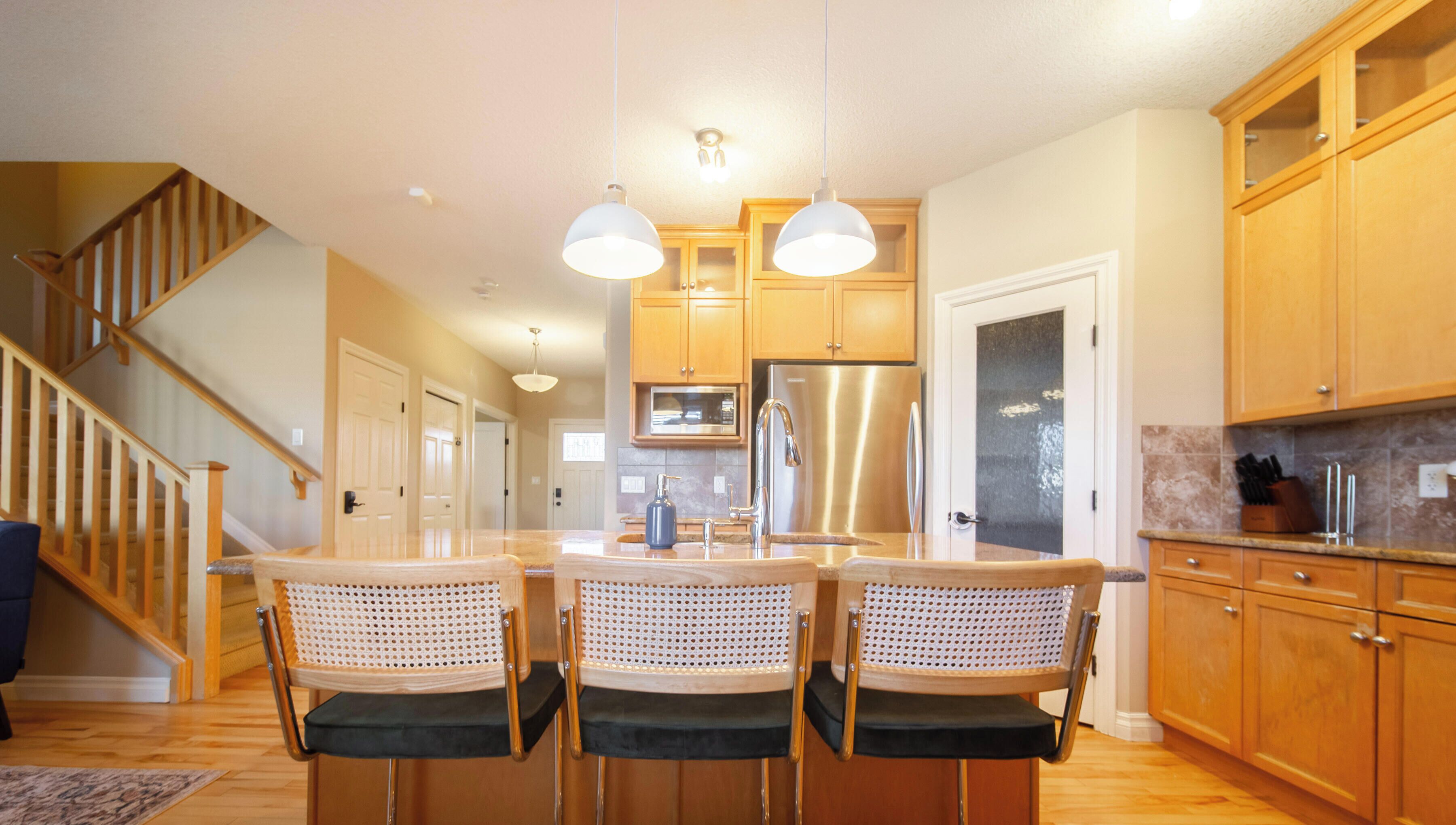 A modern kitchen with wooden cabinets, a stainless steel refrigerator, a double sink, and a dining area with three chairs.