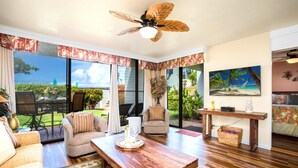 A spacious living room with a wooden coffee table, comfortable seating, a ceiling fan, and large windows offering a view of the outdoors. - Napili Point | Oceanfront 4 BR, Sleeps 16 | Car Incl. w/ 6+ Nights | NAP ML-2582 by KBM (Lahaina)