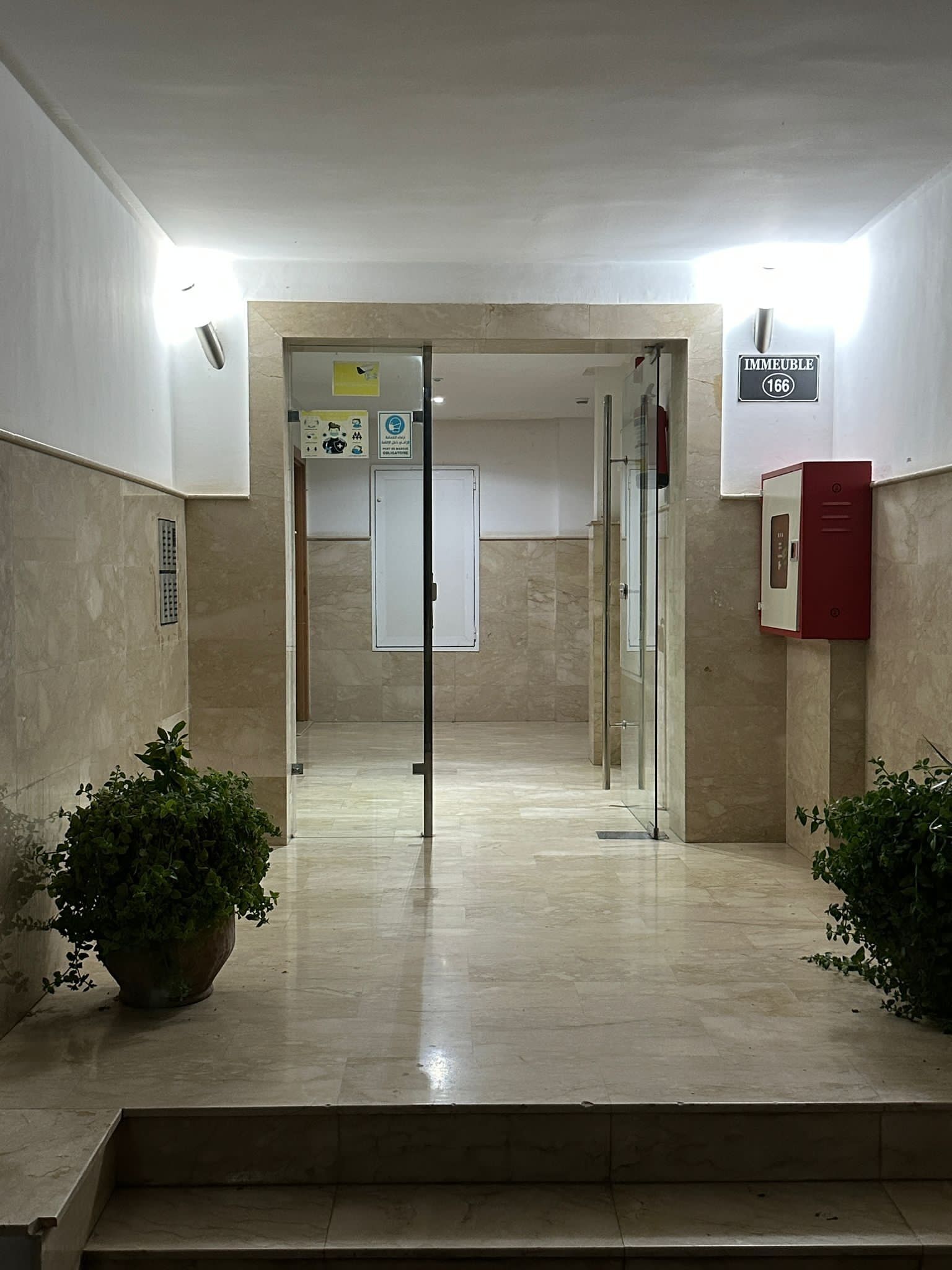 Un couloir avec une porte en verre donnant sur une pièce portant l’inscription « IMMEUBLE 166 ». De part et d’autre, on trouve des plantes en pot, et un détecteur d’incendie est fixé au mur de droite.
