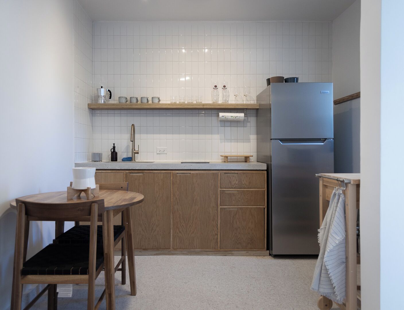 Una cocina compacta con salpicadero de azulejos blancos, armarios de madera y un refrigerador de acero inoxidable, además de una pequeña mesa de comedor con una silla.