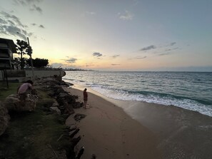 A beach with rocky terrain and a person sitting on a rock. - Garden Apartment 5 min walk to the beach. (San Juan)