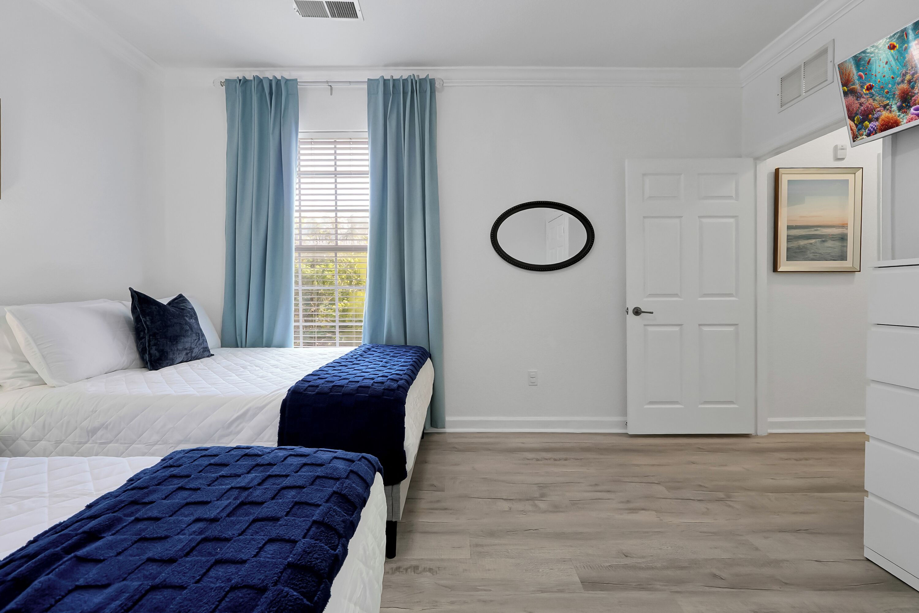 A bedroom with two beds, a window with curtains, a door, and a wall-mounted circular mirror.