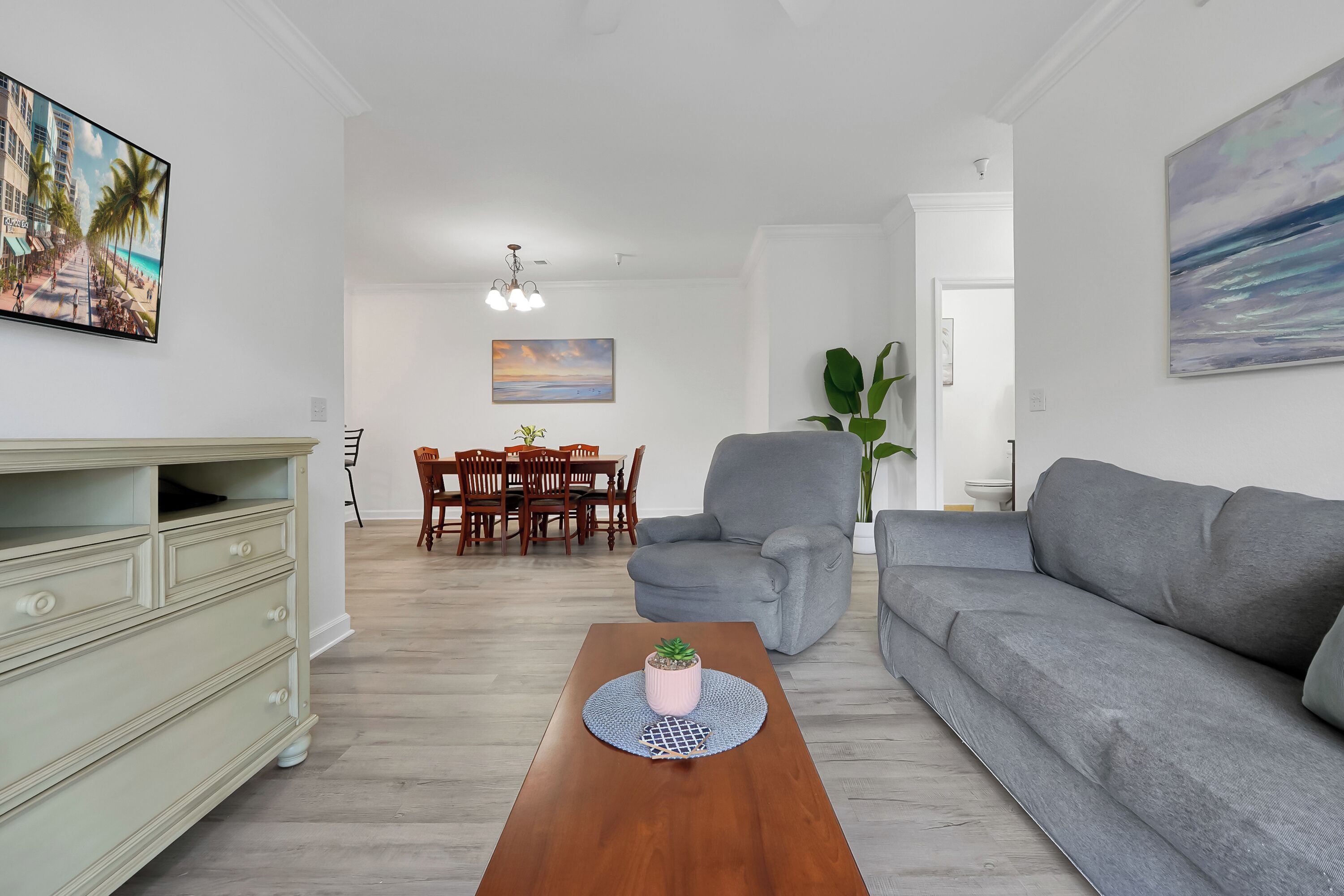 A modern living room with a grey sofa, a wooden coffee table with a plant, a dining area with a table and chairs, and a painting on the wall.