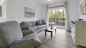 Ein Wohnzimmer mit grauem Sofa, einem Holztisch, Deckenventilator und einem großen Fenster mit Vorhängen.