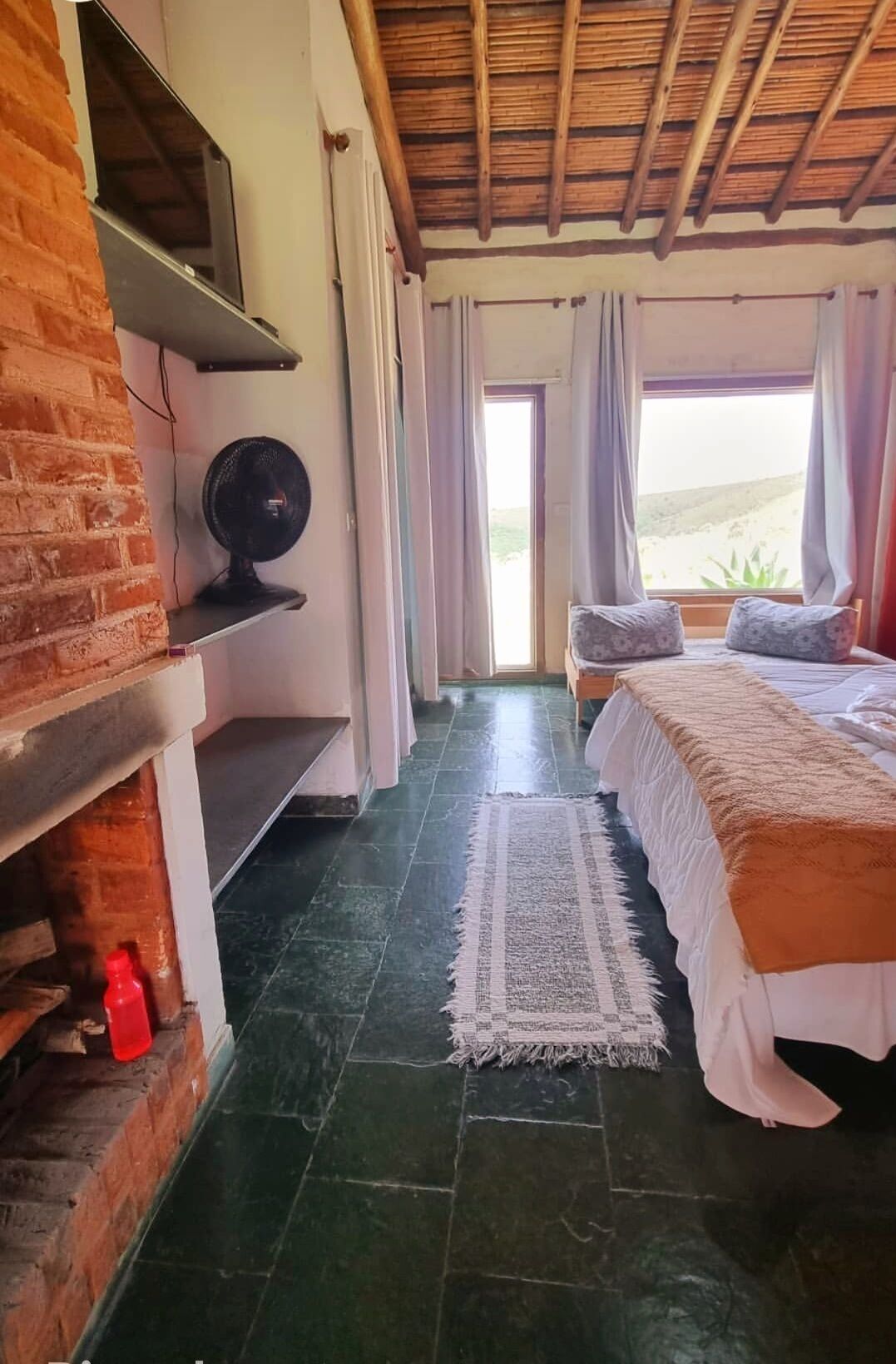 A room with a bed, a fan, a brick wall, a window with curtains, and a view outside.
