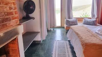 A room with a bed, a fan, a brick wall, a window with curtains, and a view outside.