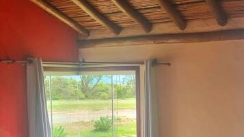 A room with two beds, a window with curtains, a red wall, and a view of greenery outside.