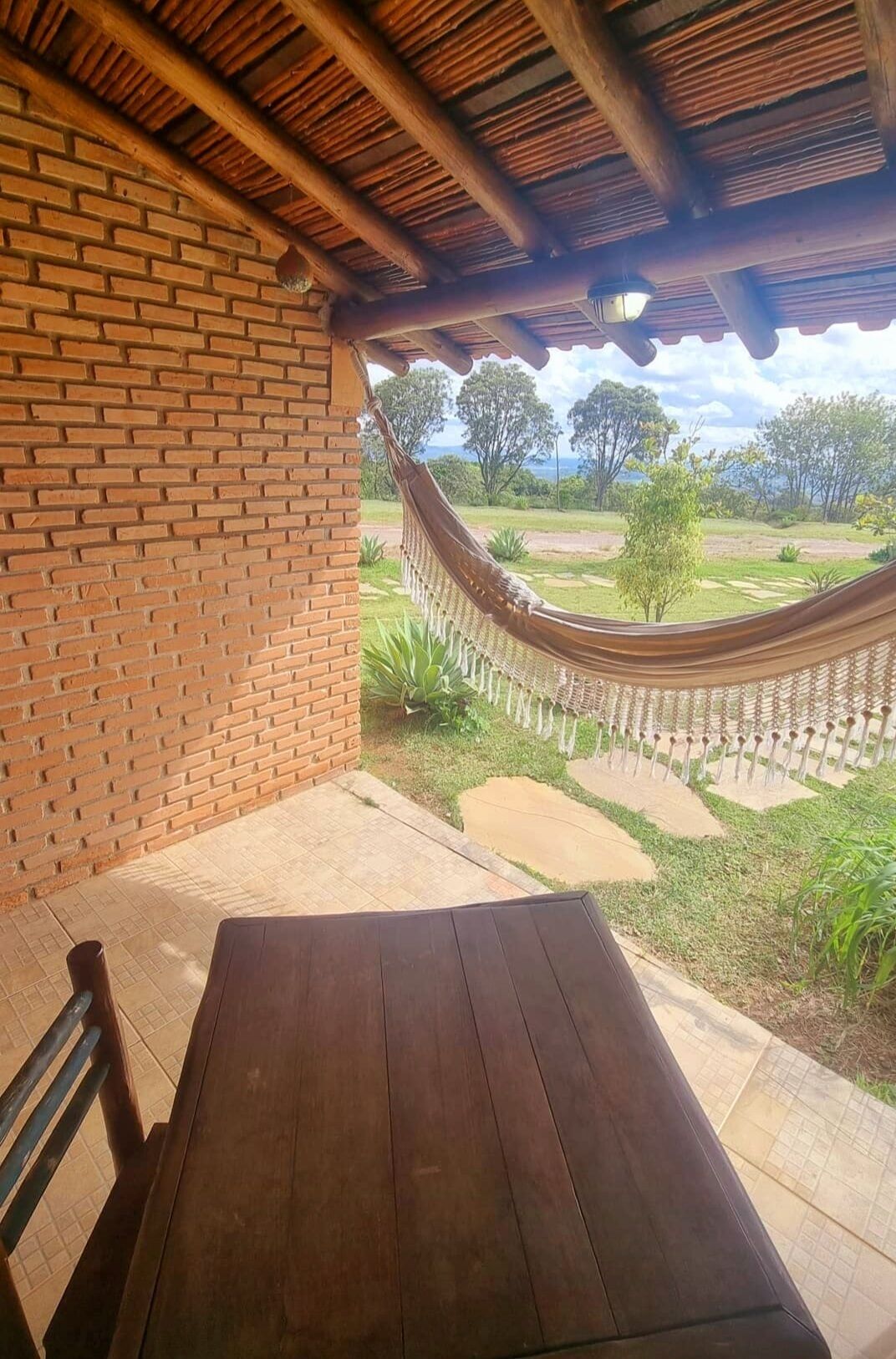 Mesa y banco de madera bajo un porche cubierto con pared de ladrillo, una hamaca y vista a un área verde con árboles.