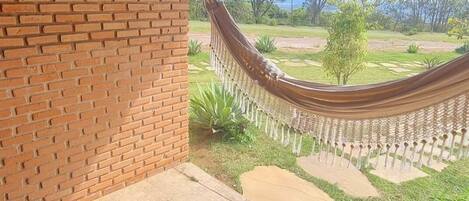 Mesa y banco de madera bajo un porche cubierto con pared de ladrillo, una hamaca y vista a un área verde con árboles.