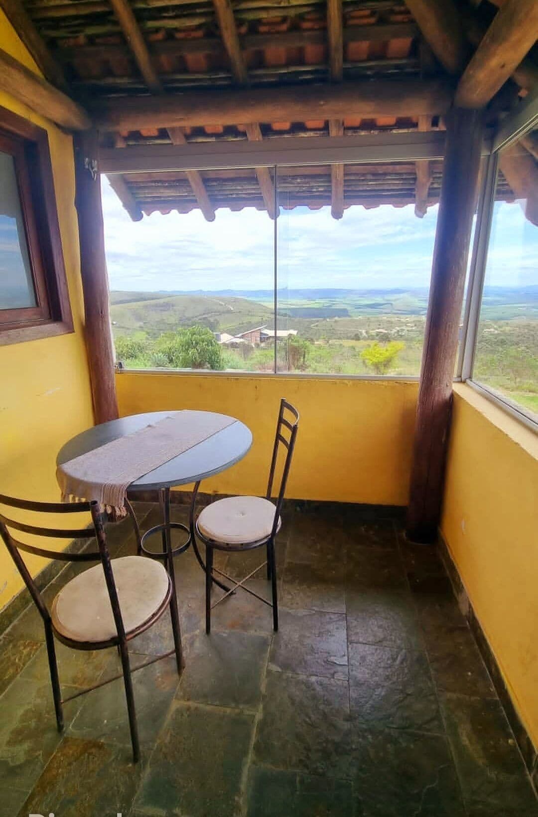 A small balcony with a table and chairs, a view of the countryside, and a wooden ceiling.