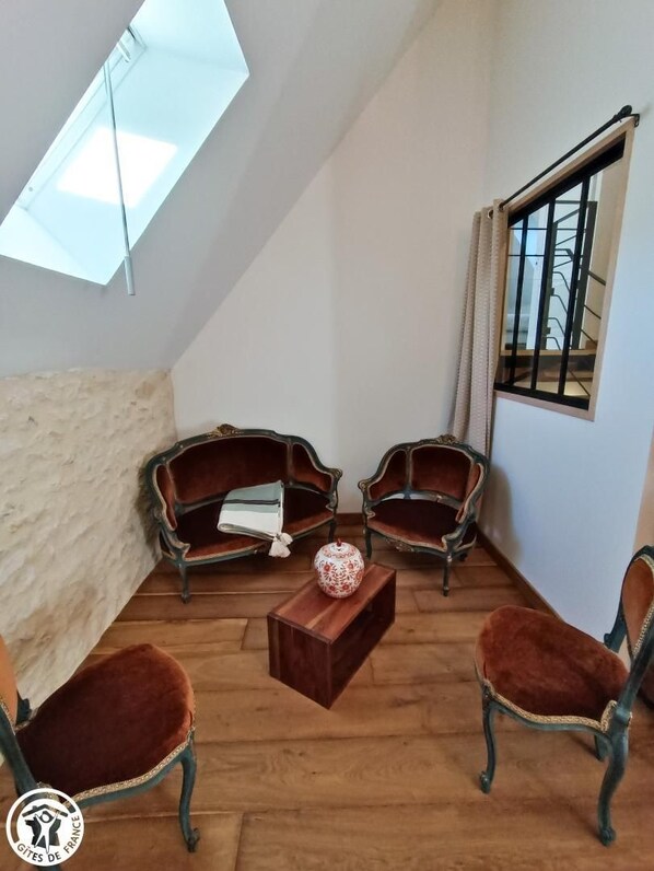 Um quarto acolhedor com duas poltronas de estilo vintage, uma mesa de centro de madeira e um pequeno vaso decorativo. Um clarabóia oferece luz natural, e uma janela dá vista para uma escadaria.