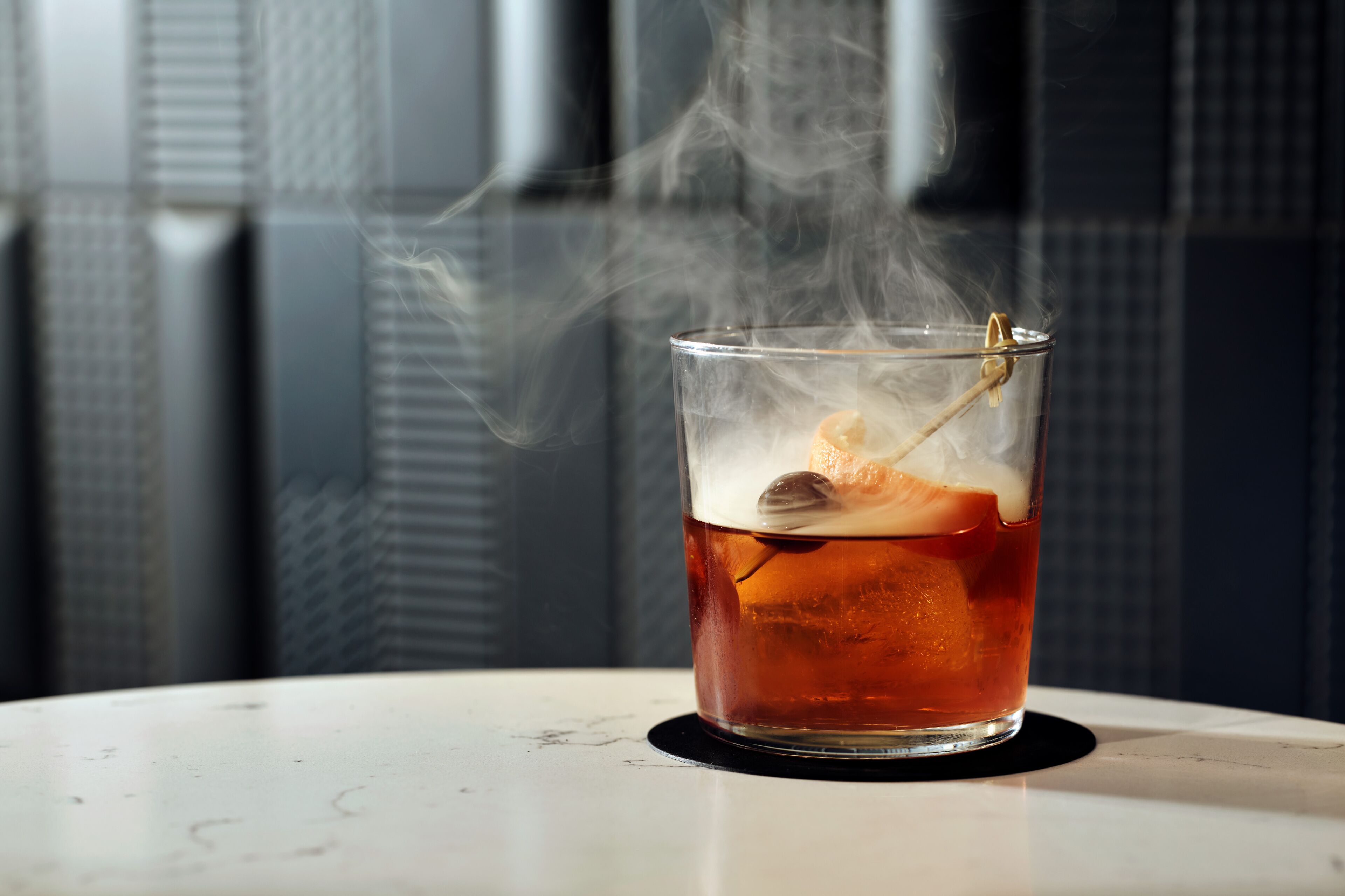 Un verre contenant un liquide ambré, avec des glaçons et une garniture, dont s'échappe de la vapeur, posé sur une surface blanche, le tout sur un fond à motifs.