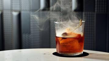 Un vaso de líquido ámbar con hielo y un adorno, vapor saliendo de él, sobre una superficie blanca con un fondo estampado.