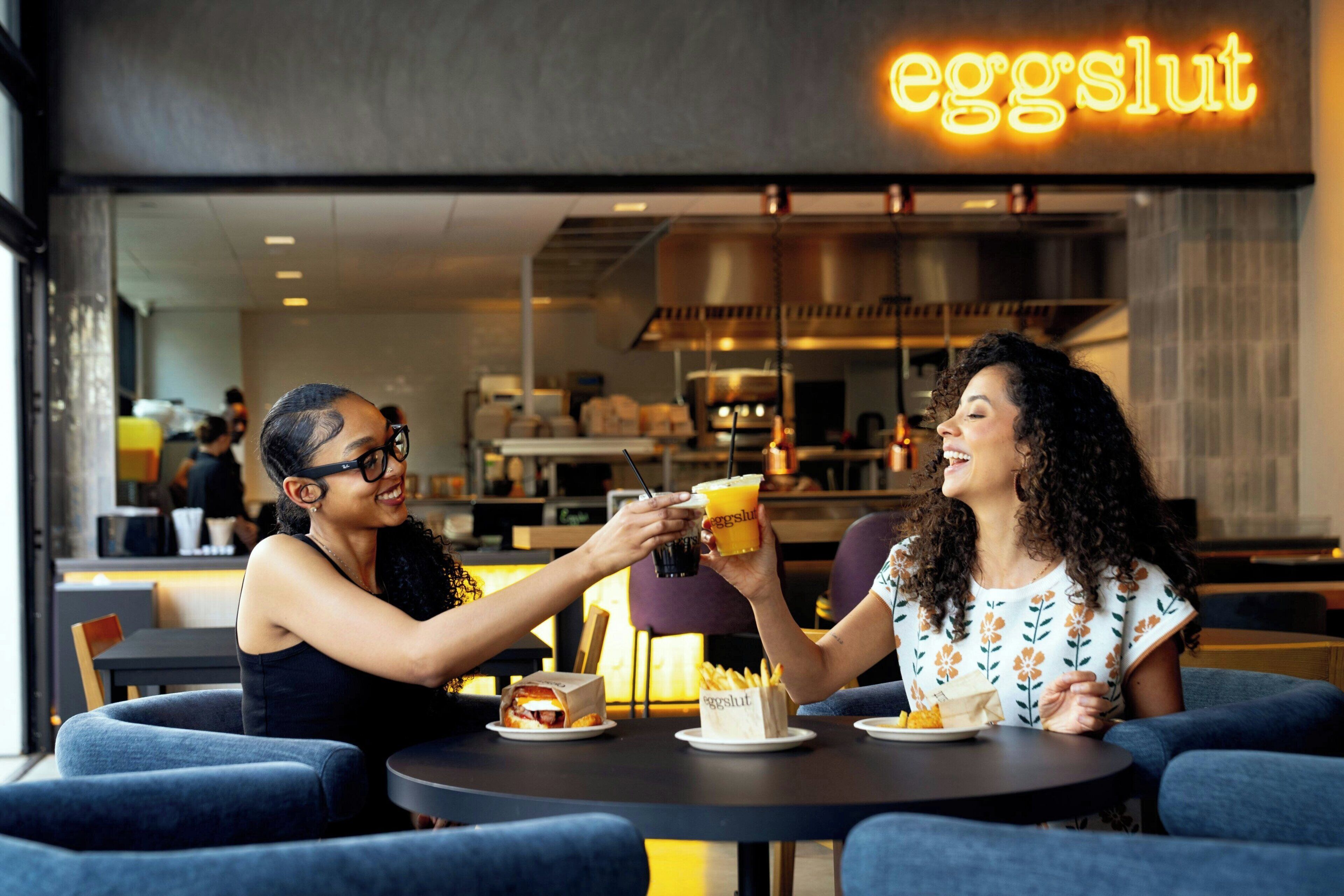 Two people toasting with drinks at a table in a cafe with a neon sign reading 'eggshut' in the background.