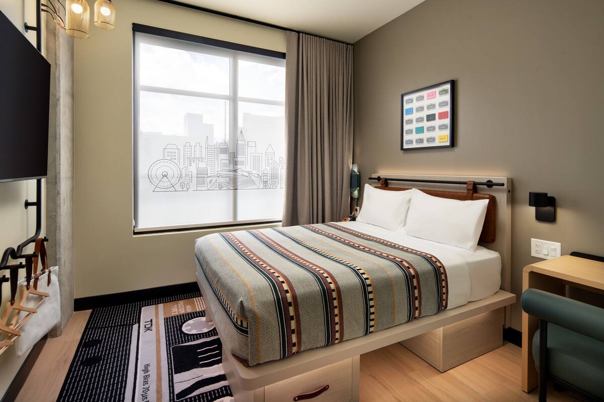 A hotel room with a bed, a desk, a chair, a window with a cityscape view, and a wall-mounted picture.