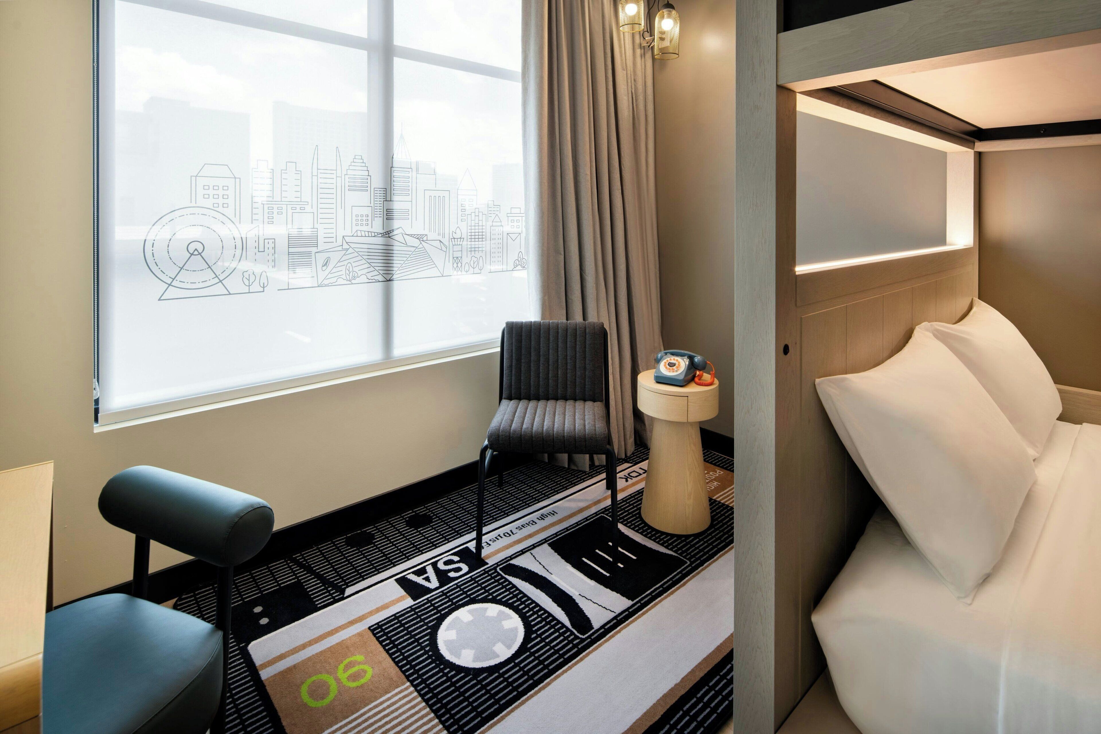 Hotelkamer met uitzicht op de stad, een bed, een stoel, een klein tafeltje en een decoratief object op het nachtkastje.