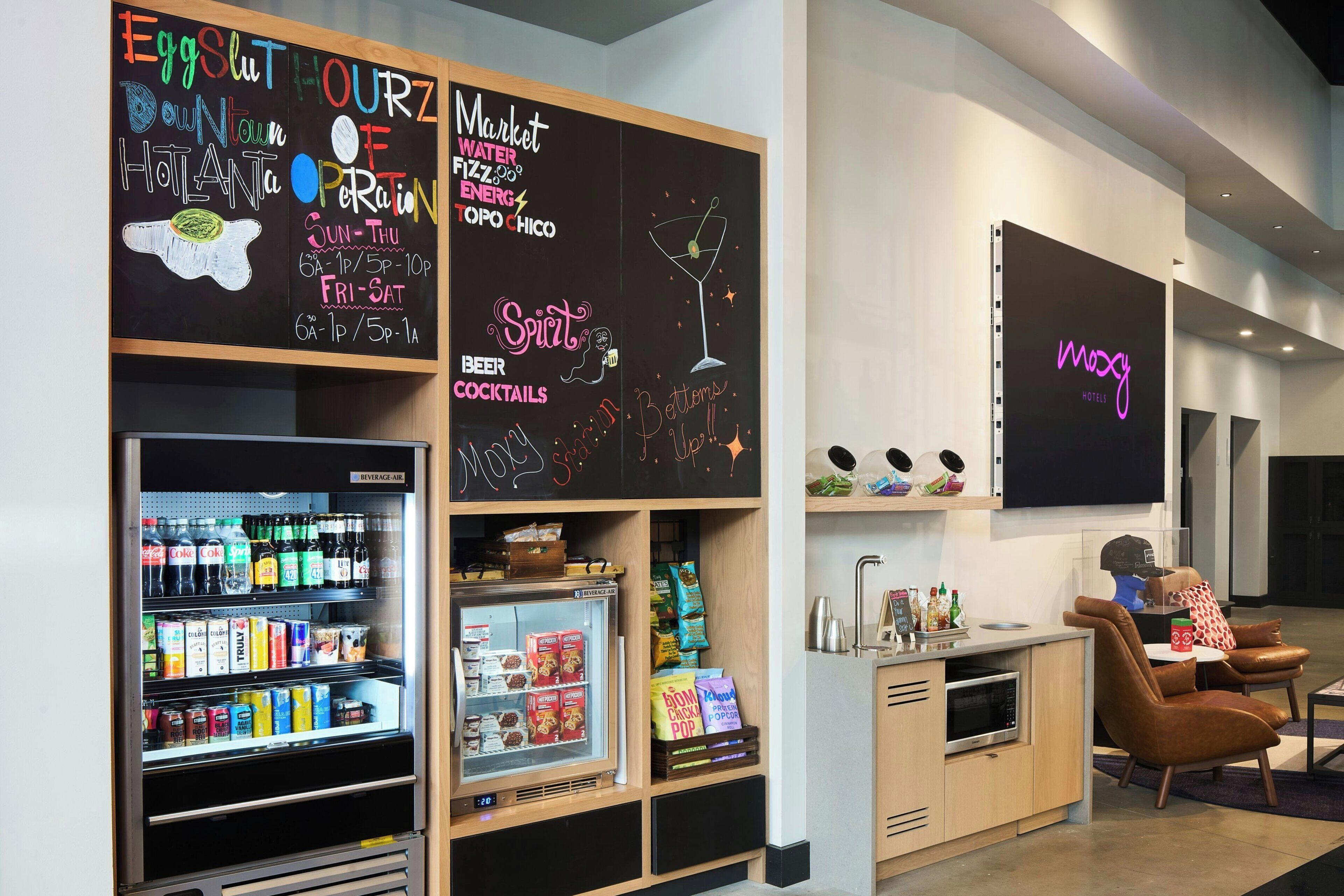 A hotel lobby with a refrigerator stocked with beverages, a minibar, and a chalkboard with the hotel's name and amenities.
