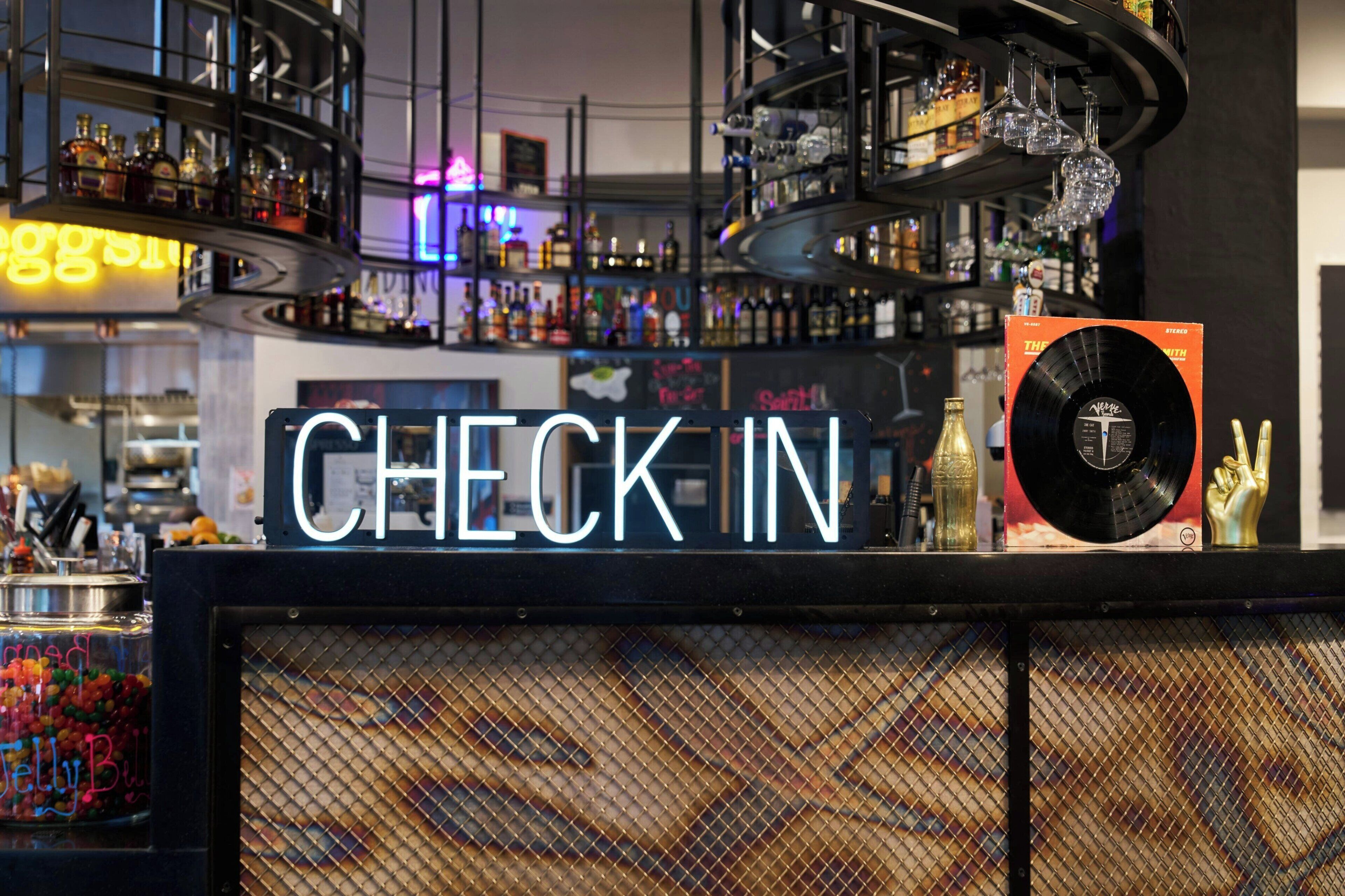 Zona de bar con un letrero de neón que dice 'CHECK IN', un disco de vinilo y varios botellas y vasos.