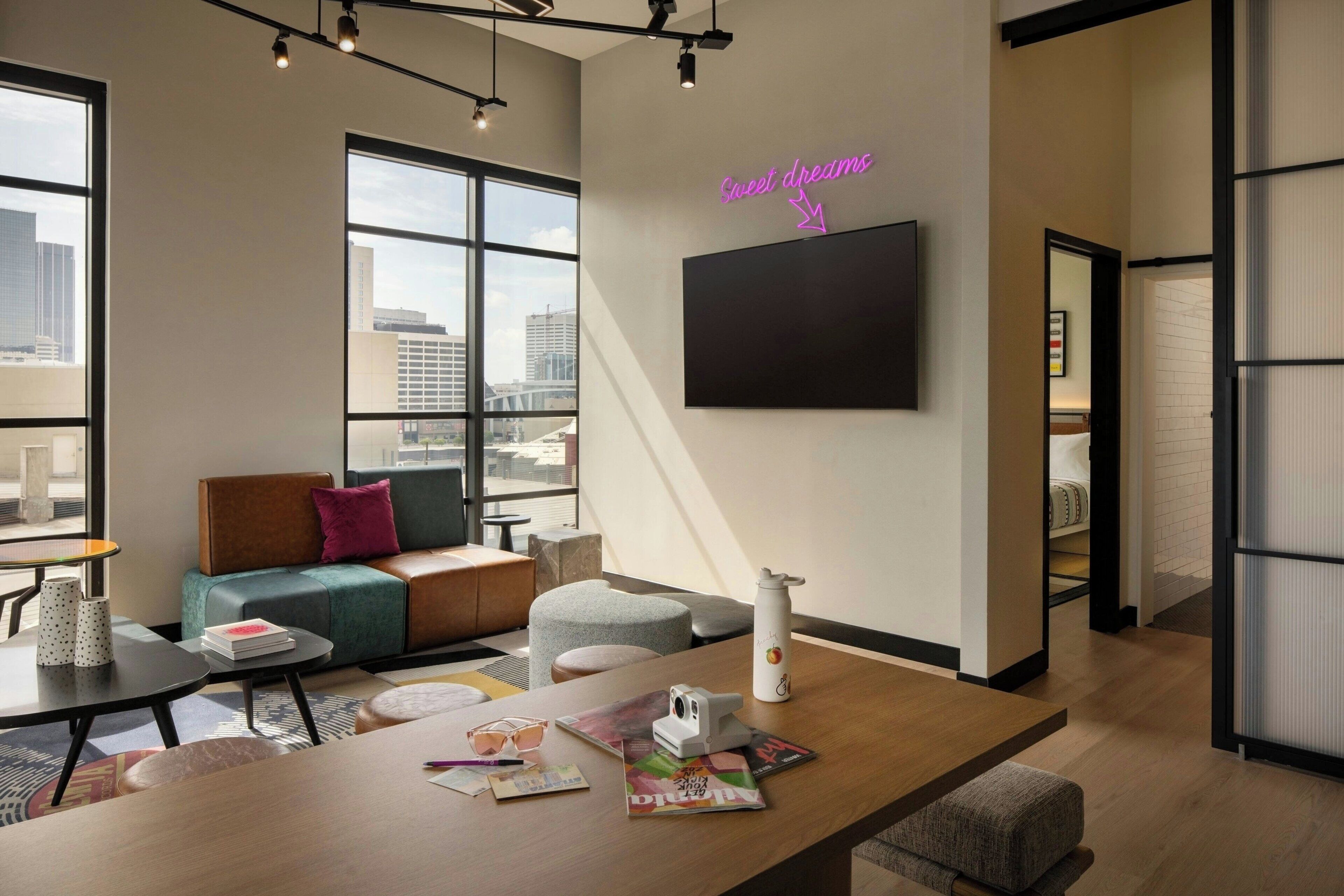 A modern hotel room with a large window, a flat-screen TV, a neon sign, a sofa, a coffee table, and a dining table.