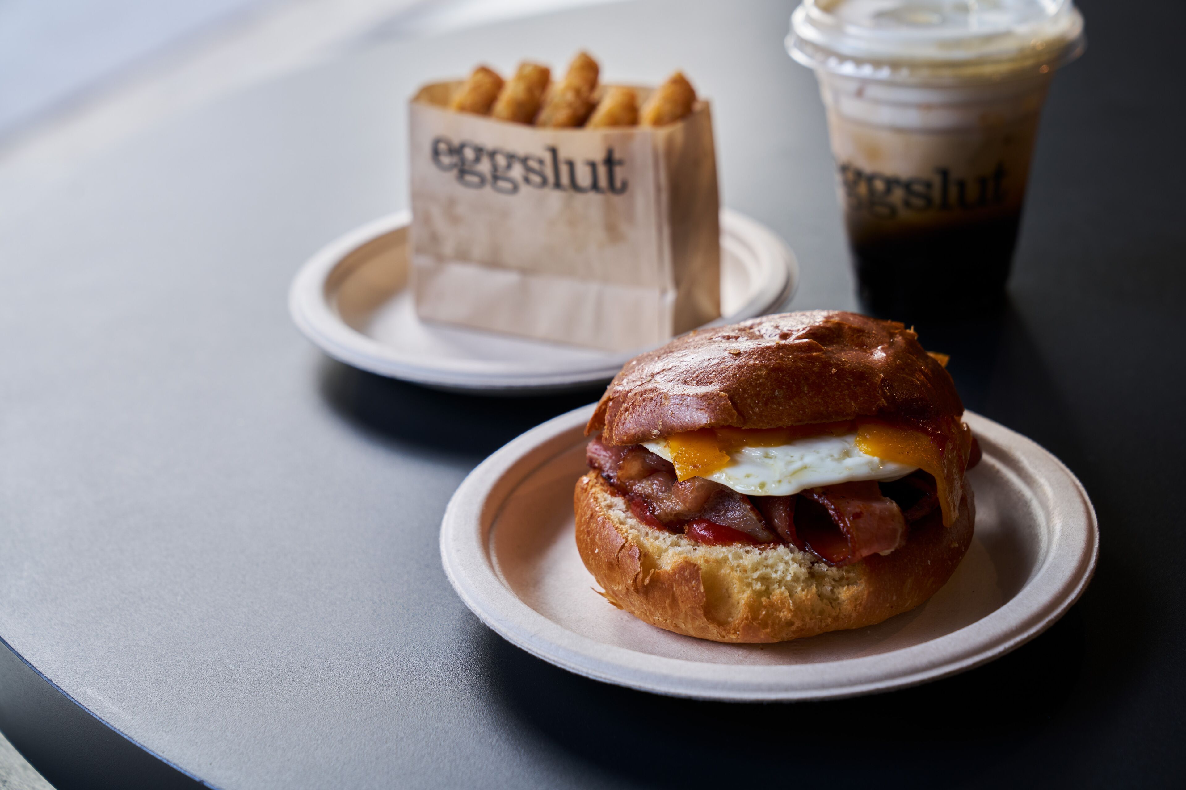 Un sandwich sur une assiette, un sac en papier avec l’inscription « eggslut », et une tasse de café glacé.