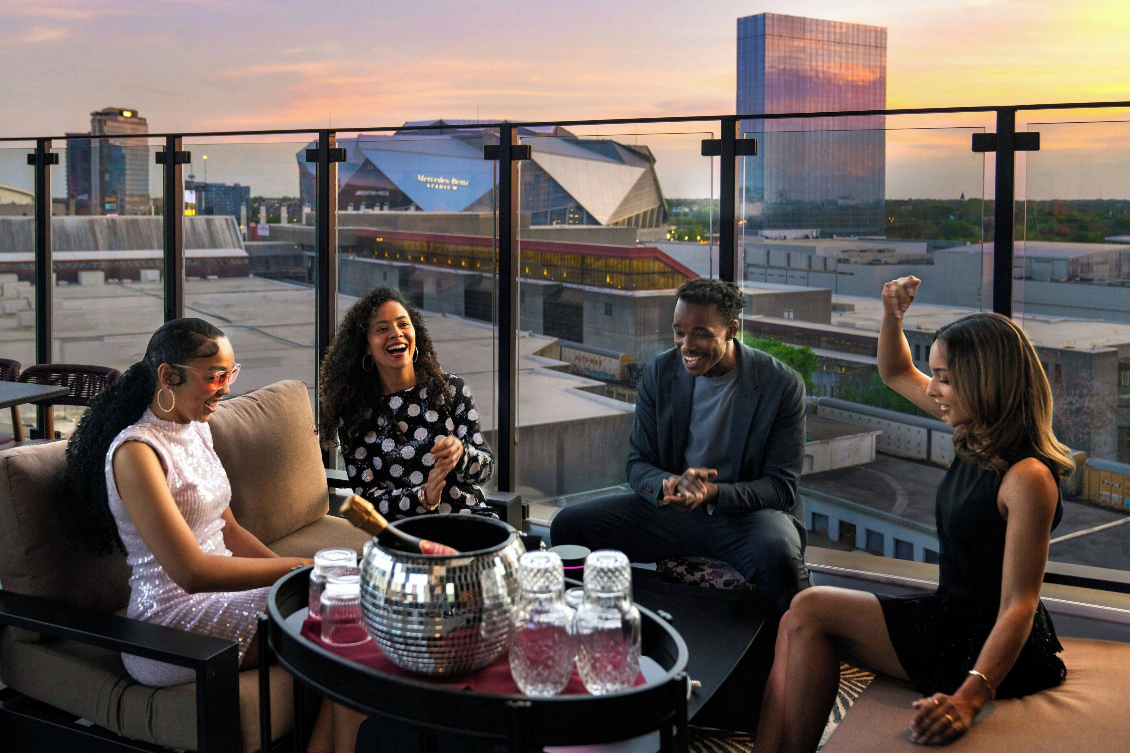 A group of people seated on a rooftop terrace with a cityscape view, including a modern building and a stadium.