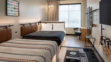 Habitación de hotel con dos camas, un escritorio, una silla, una ventana con cortinas, un televisor montado y un perchero con bolsos.