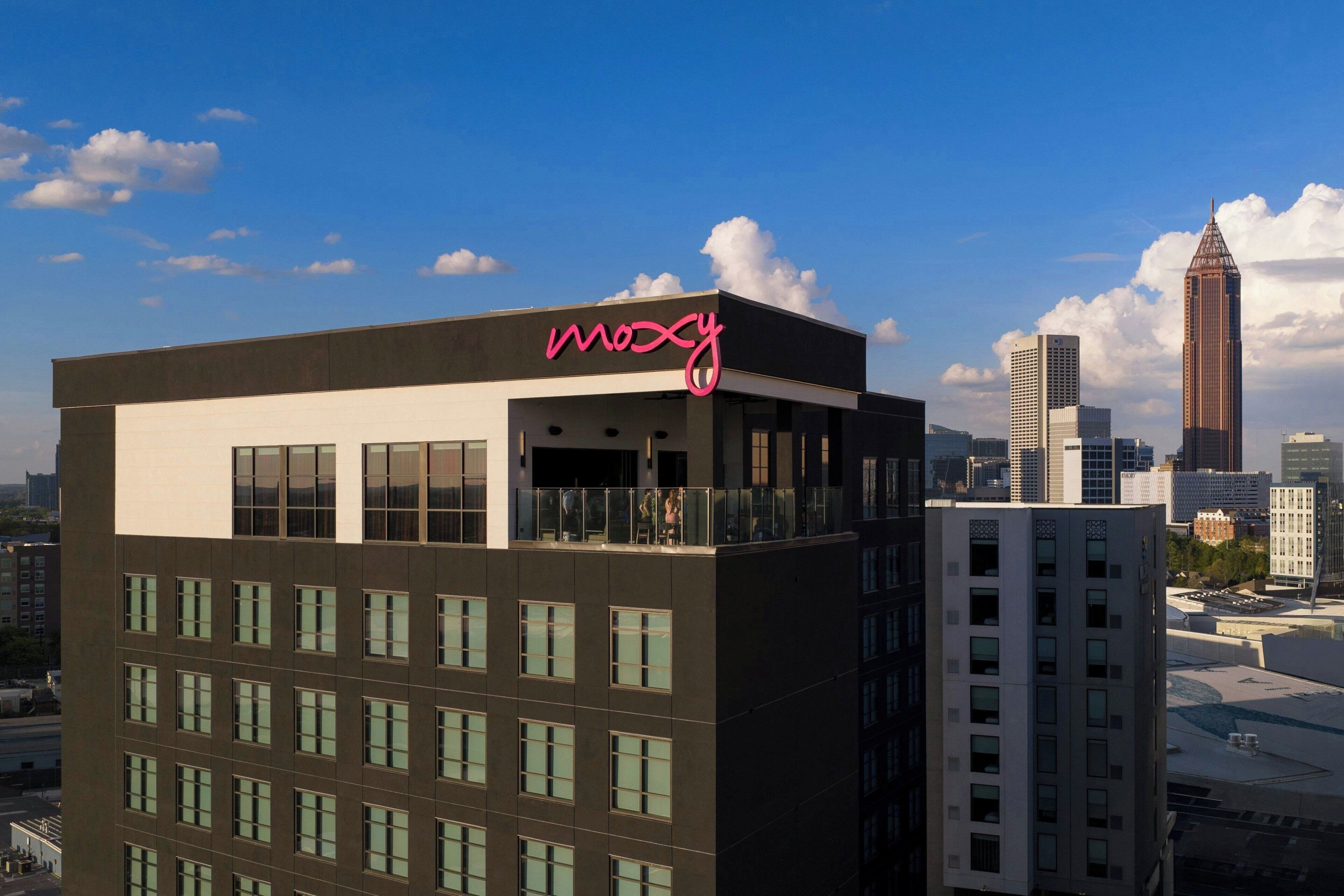 Un bâtiment moderne, surmonté d’un grand panneau lumineux portant l’inscription « Moxy », entouré d’autres immeubles de grande hauteur et se dressant sous un ciel dégagé.