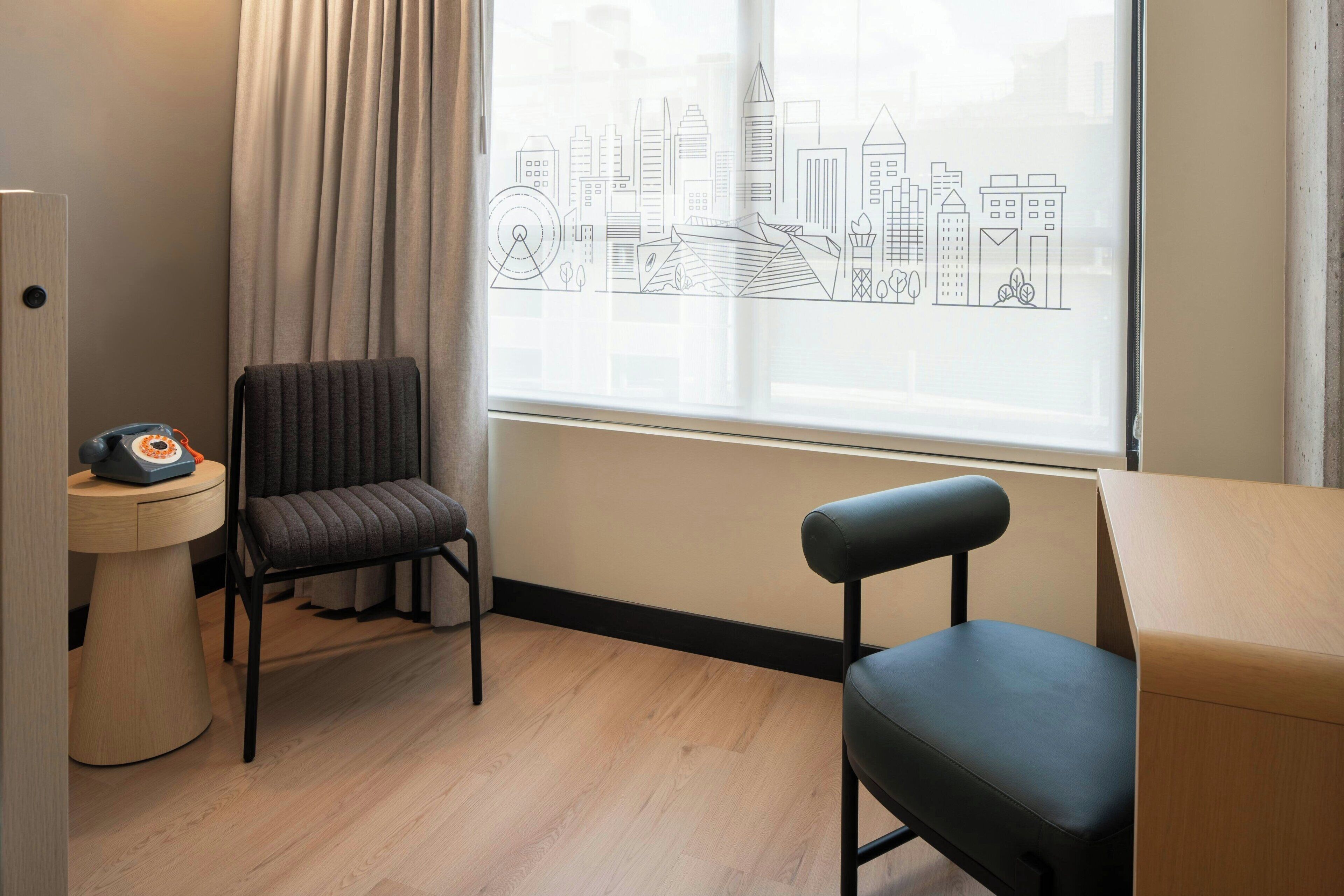 A room with a cityscape view, a chair, a small table with a telephone, and a window with curtains.