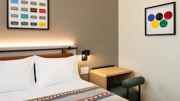 A hotel room with a bed, bedside table, chair, wall-mounted artwork, and a wall socket.