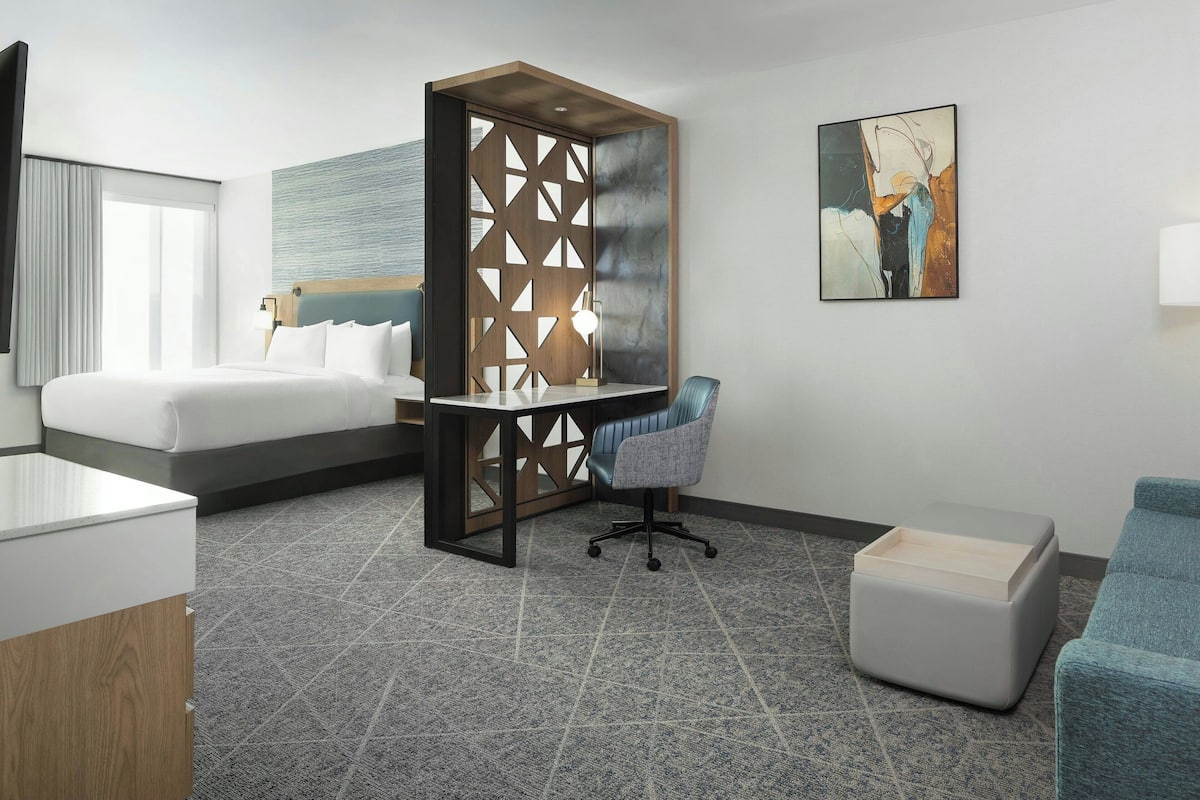 A hotel room with a bed, desk, chair, ottoman, and artwork on the wall.