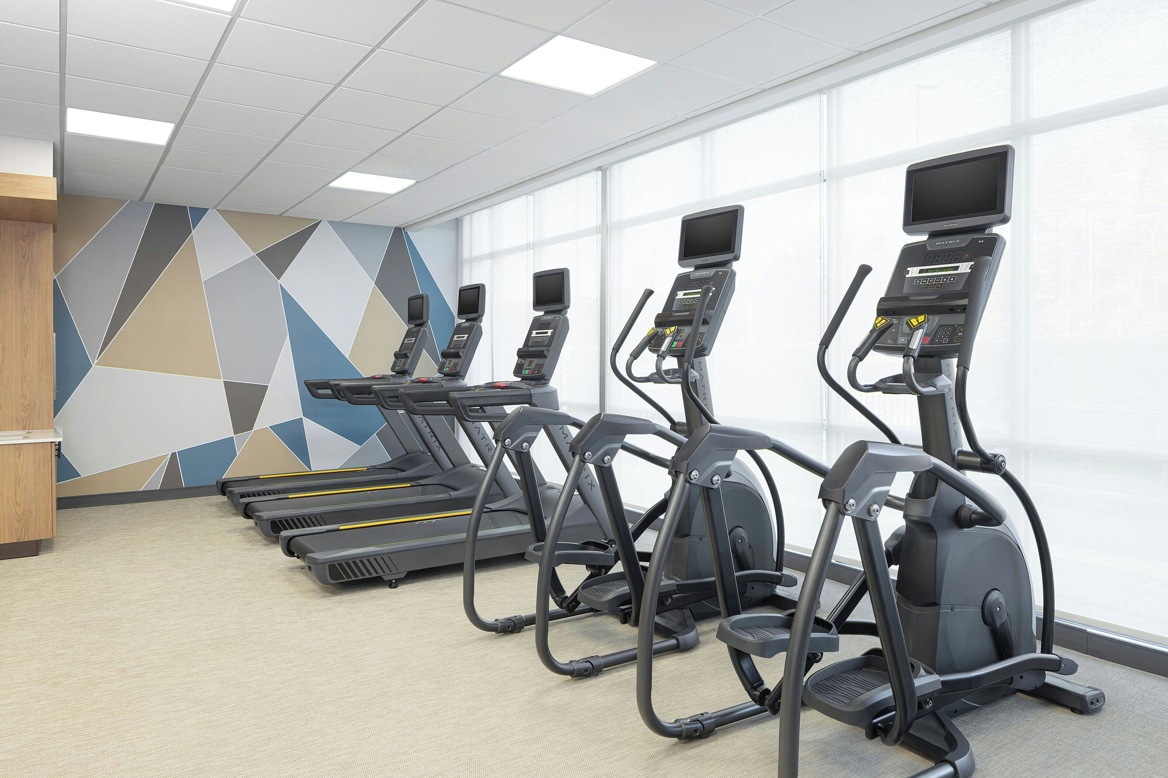 A row of treadmills and elliptical machines in a gym with a geometric wall design and large windows.