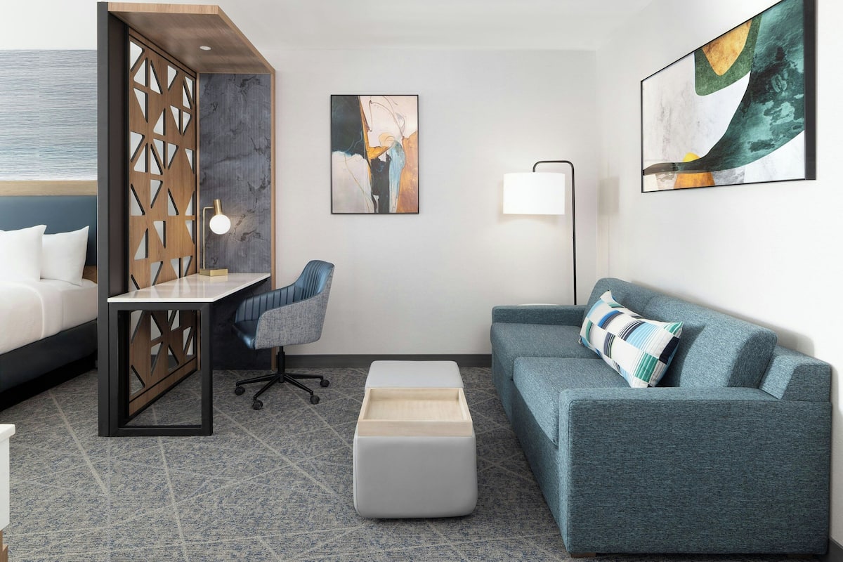 A modern hotel room with a bed, a desk, a sofa, and a chair. There are abstract paintings on the walls and a window with a view.