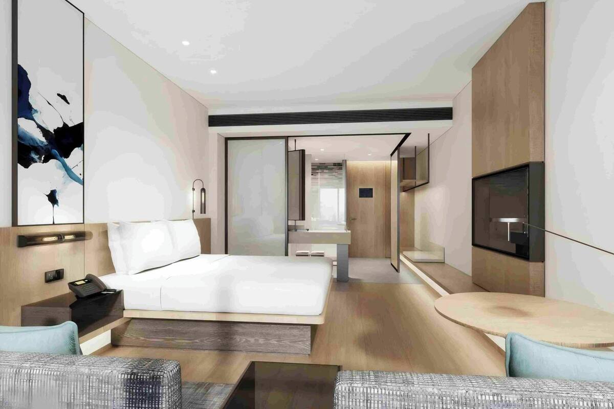 Una habitación de hotel moderna con una cama grande, un área de descanso con una mesa redonda, un televisor y un baño con espejo y lavamanos.