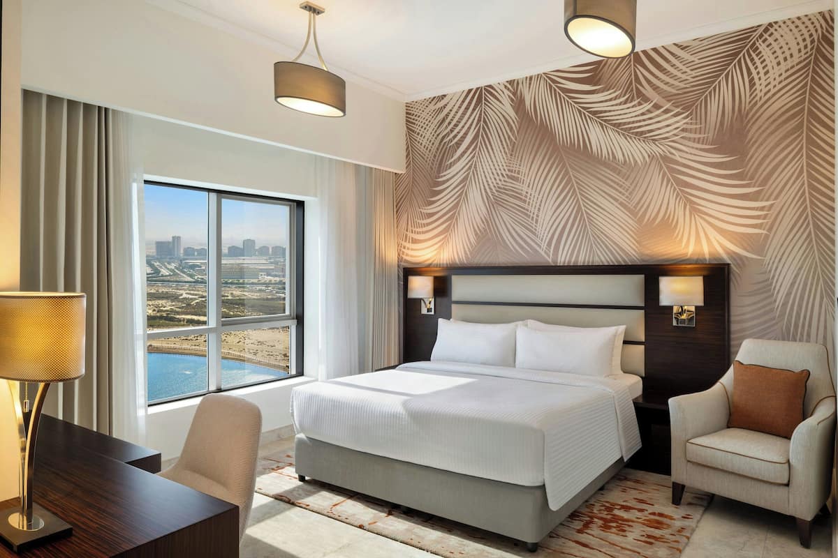 A hotel room with a large bed, a desk, a chair, and a view of a cityscape through large windows.