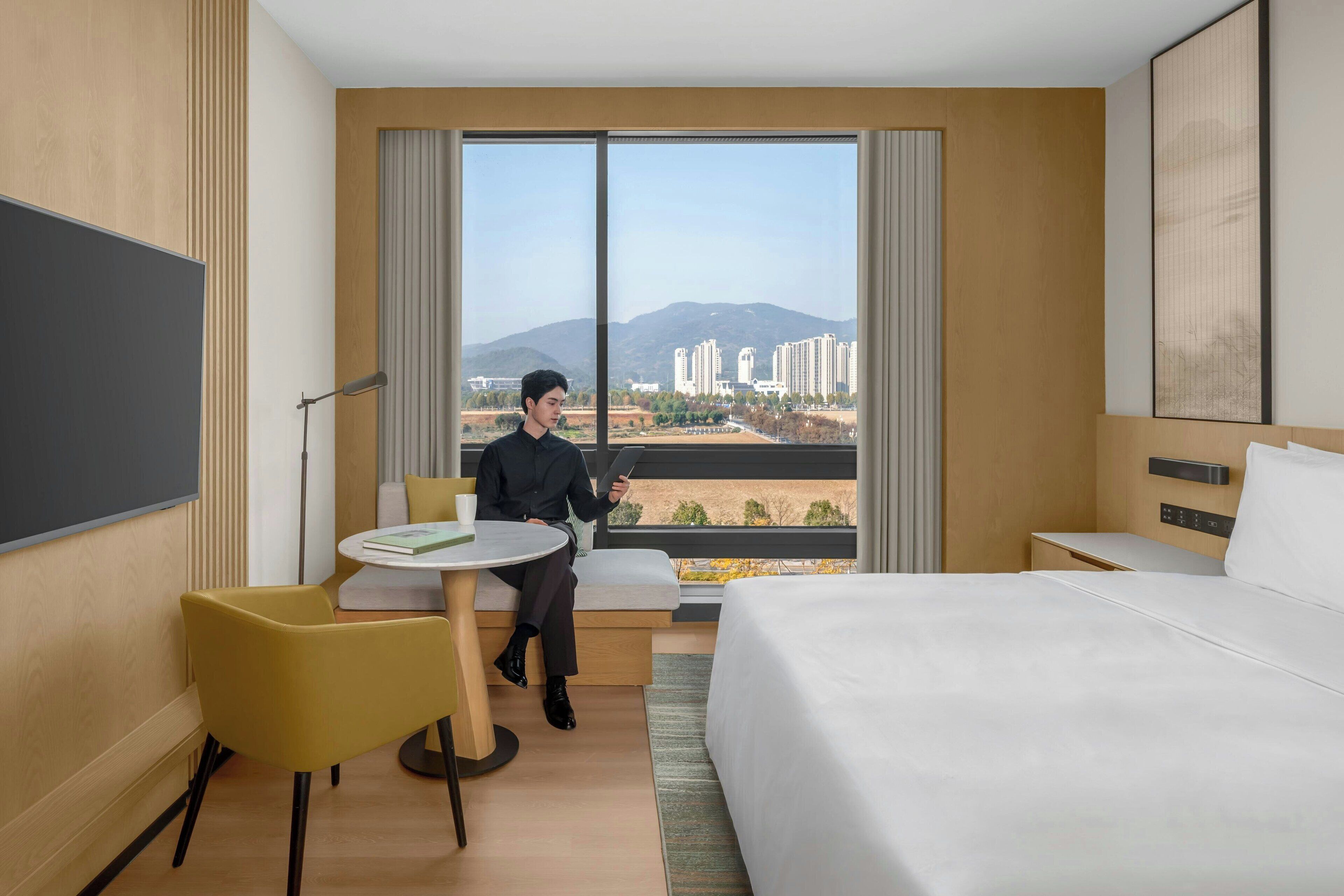 A hotel room with a large bed, a small round table, a yellow chair, and a view of buildings and mountains through large windows.