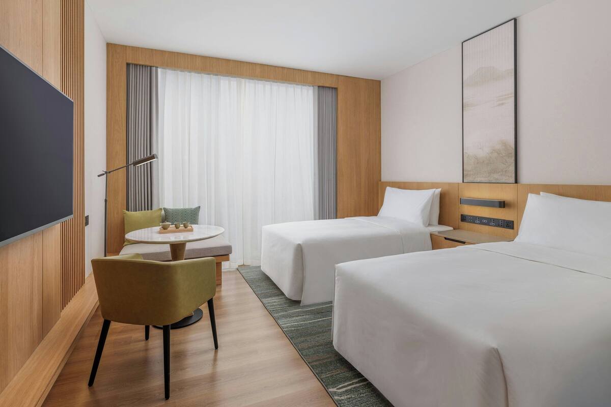 Une chambre d’hôtel avec deux lits, une télévision, une petite table avec des chaises et une grande fenêtre avec des rideaux.