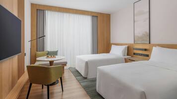 Habitación de hotel con dos camas, televisión, una mesita con sillas y un ventanal grande con cortinas.