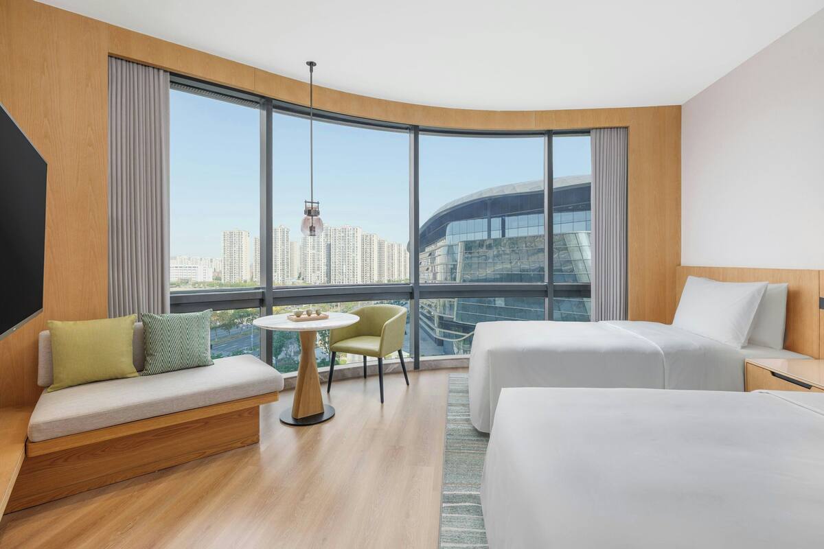 Une chambre d’hôtel moderne avec une fenêtre incurvée, un canapé, deux lits et une petite table.