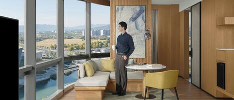 A modern hotel room with a large window offering a city view, a sofa, a dining table, and a minimalist decor.