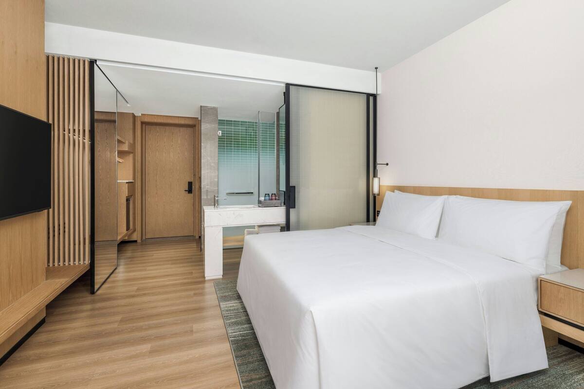 Une chambre d’hôtel moderne, dotée d’un grand lit, d’une table de chevet, d’une télévision, d’une armoire et d’une salle de bain visible à travers une porte coulissante.