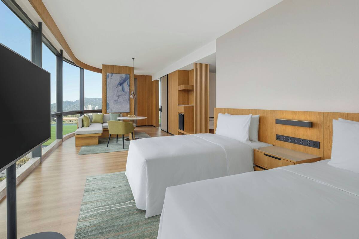 Une chambre d’hôtel moderne dotée de deux lits, d’un téléviseur à écran plat, d’un coin salon avec vue et d’une grande fenêtre.