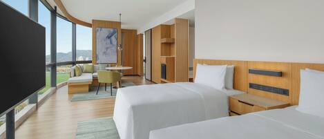 A modern hotel room with two beds, a flat-screen TV, a seating area with a view, and a large window.