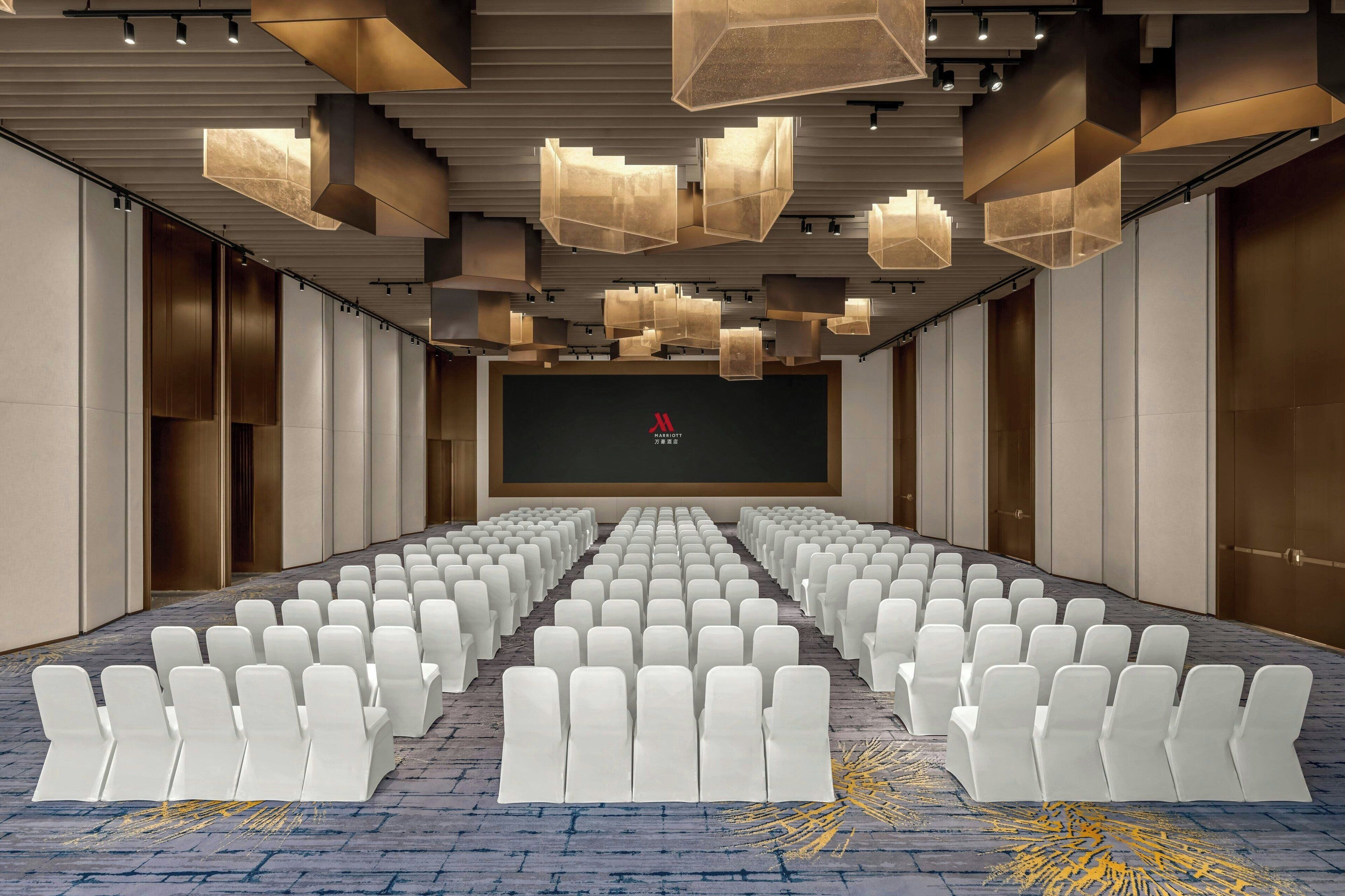 A conference room with rows of white chairs, a large screen at the front, and modern geometric ceiling lights.