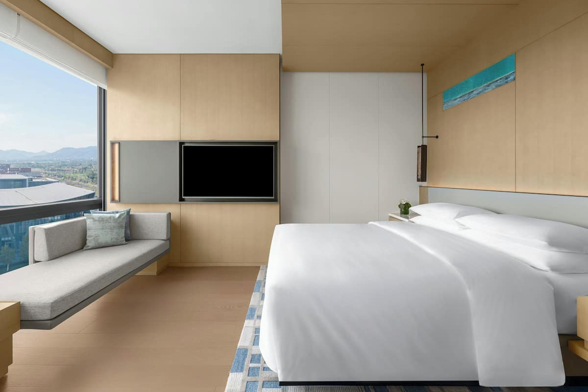 Ein modernes Hotelzimmer mit einem großen Bett, einer integrierten Sitzbank mit Kissen, einem Flachbildfernseher und Blick auf die Stadt.