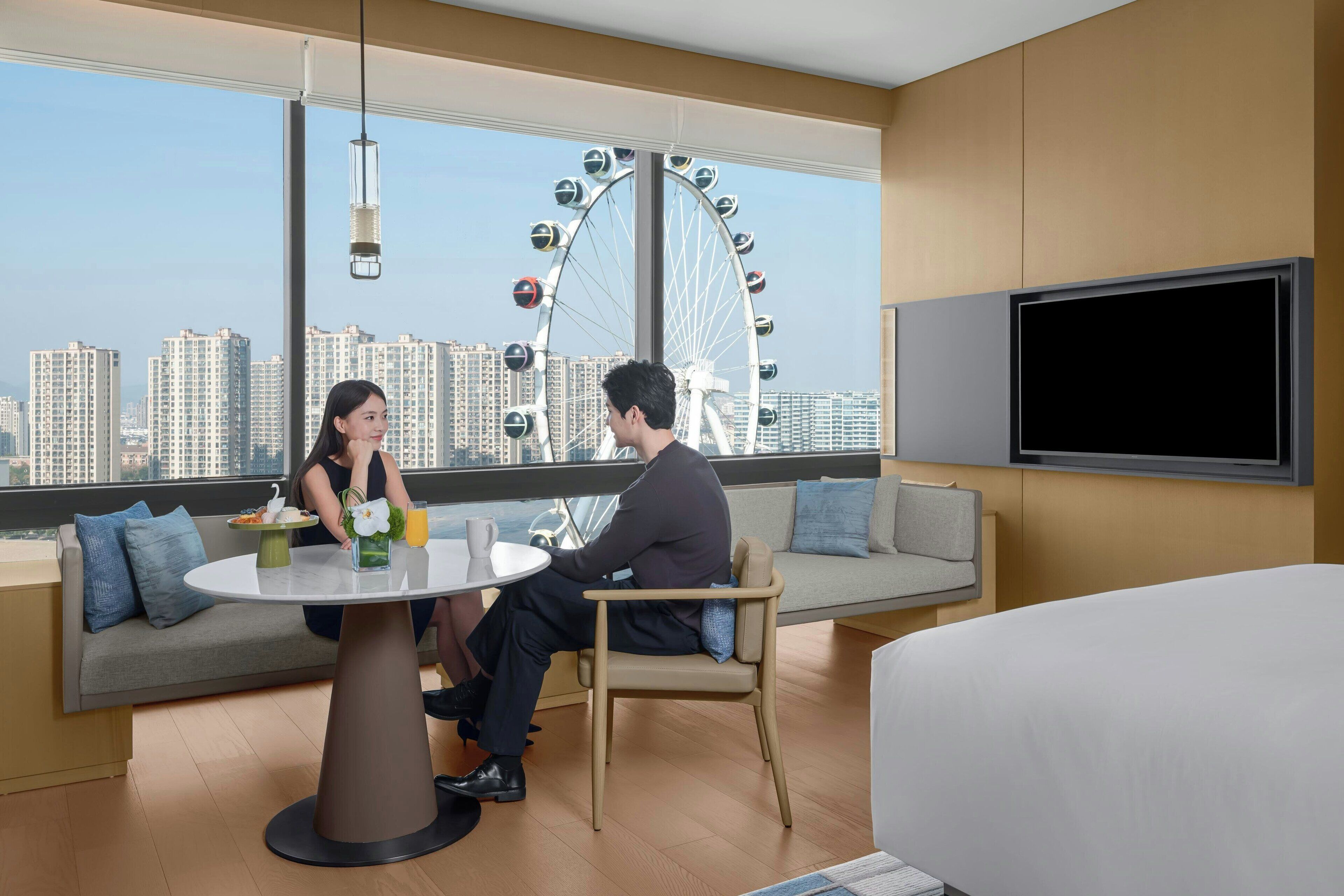 Une chambre d’hôtel moderne dotée d’une grande fenêtre offrant une vue sur une grande roue et des immeubles de grande hauteur. La chambre comprend une table ronde, un canapé et un lit.
