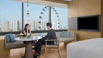 Une chambre d’hôtel moderne dotée d’une grande fenêtre offrant une vue sur une grande roue et des immeubles de grande hauteur. La chambre comprend une table ronde, un canapé et un lit.