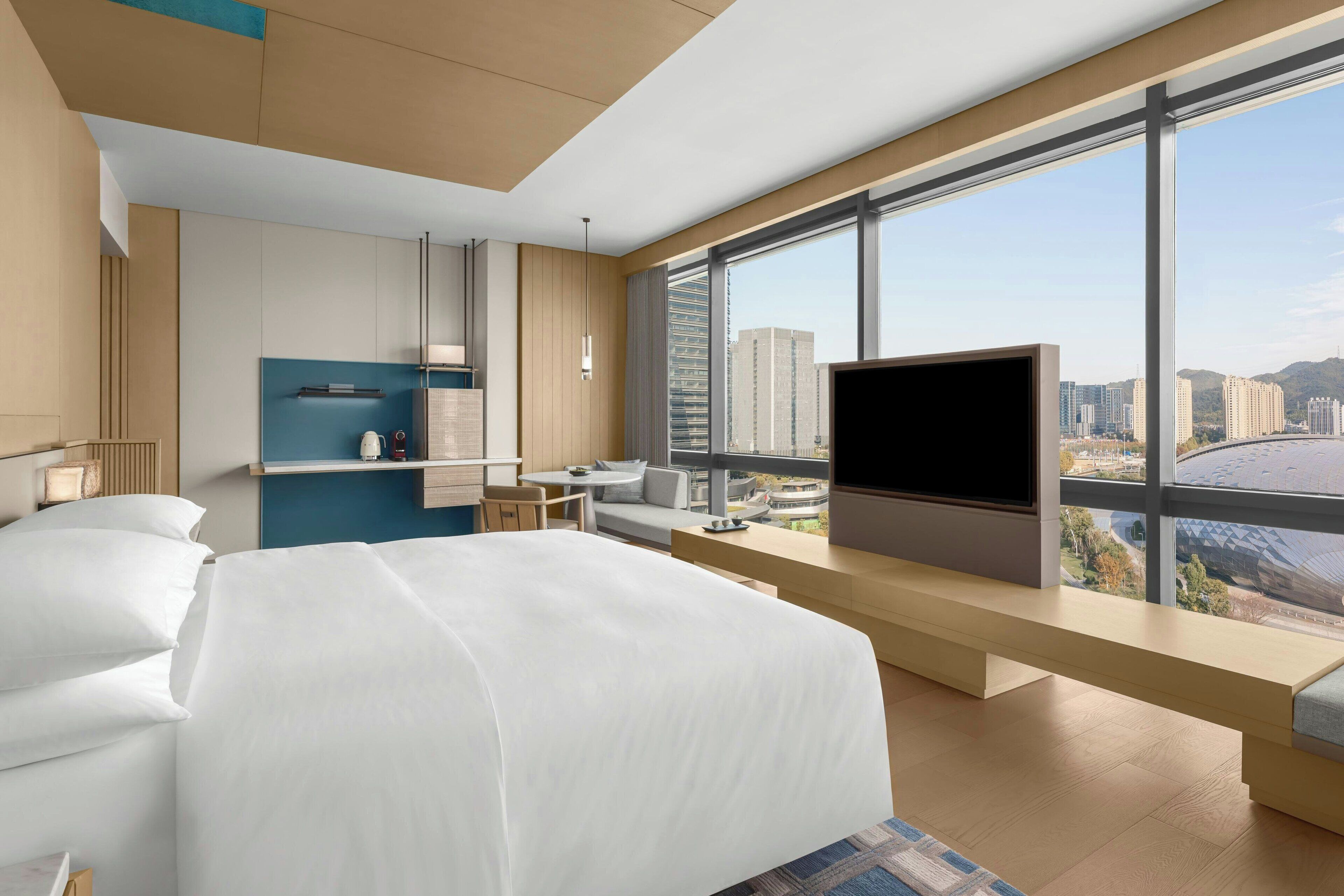 Ett modernt hotellrum med en stor säng, en TV, ett skrivbord och utsikt över stadsbilden genom stora fönster.